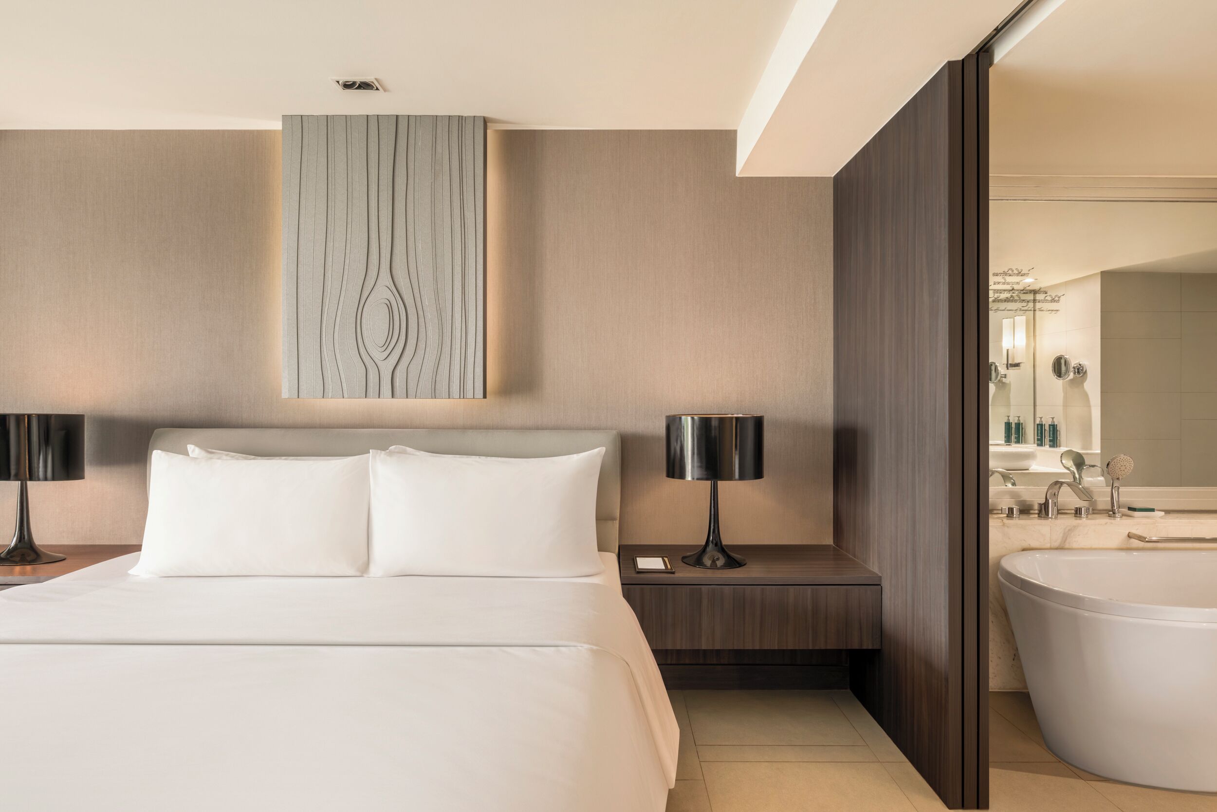 A modern hotel room with a large bed, bedside lamps, a wall-mounted artwork, a bathroom with a bathtub, and a large mirror.