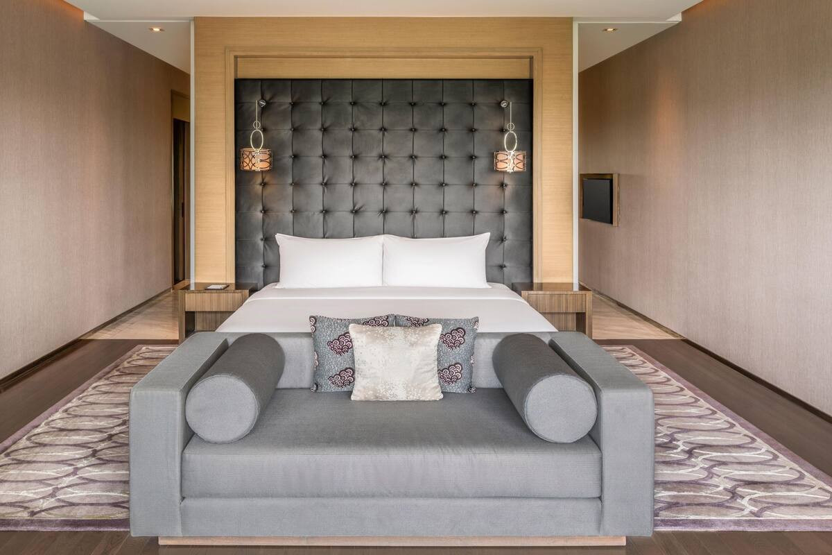 A hotel room with a large bed, a grey sofa, a wooden headboard, two bedside lamps, and a flat-screen TV.