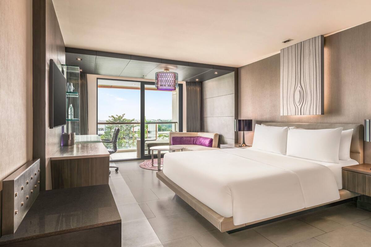 A modern hotel room with a large bed, a desk, a chair, a balcony view, and a large artwork on the wall.
