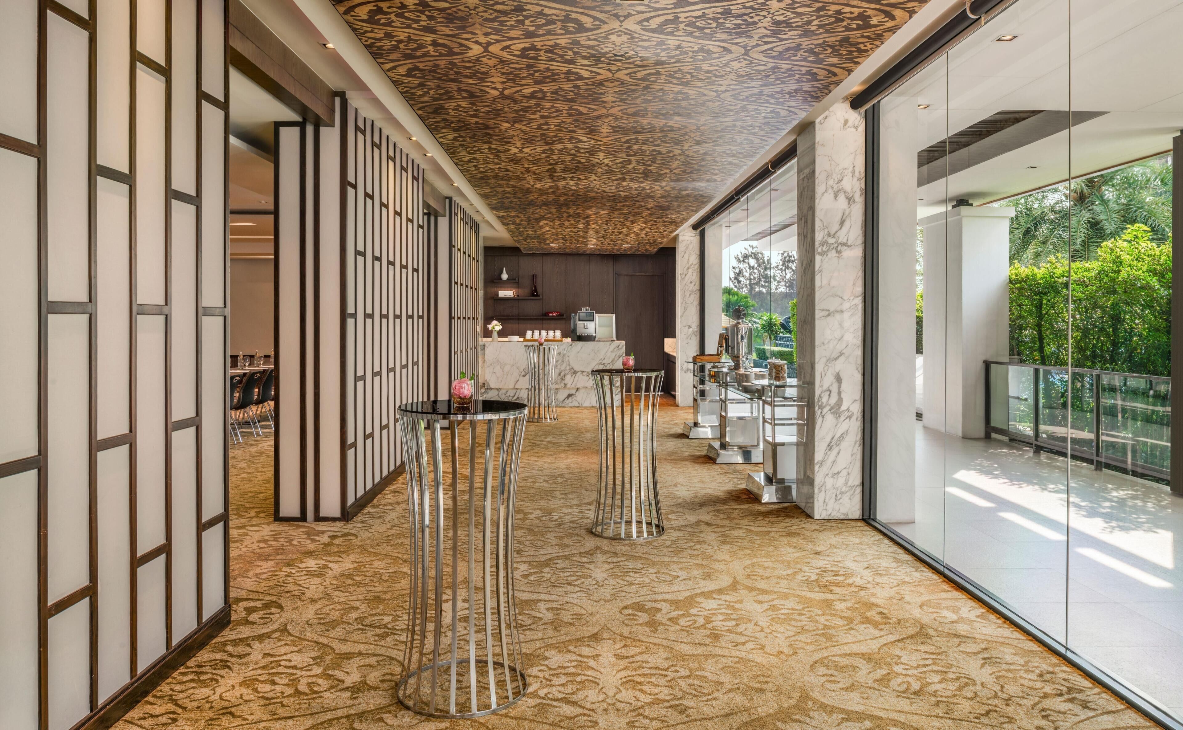 A corridor with patterned carpet, metal stools, a reception desk, and a view of greenery outside.