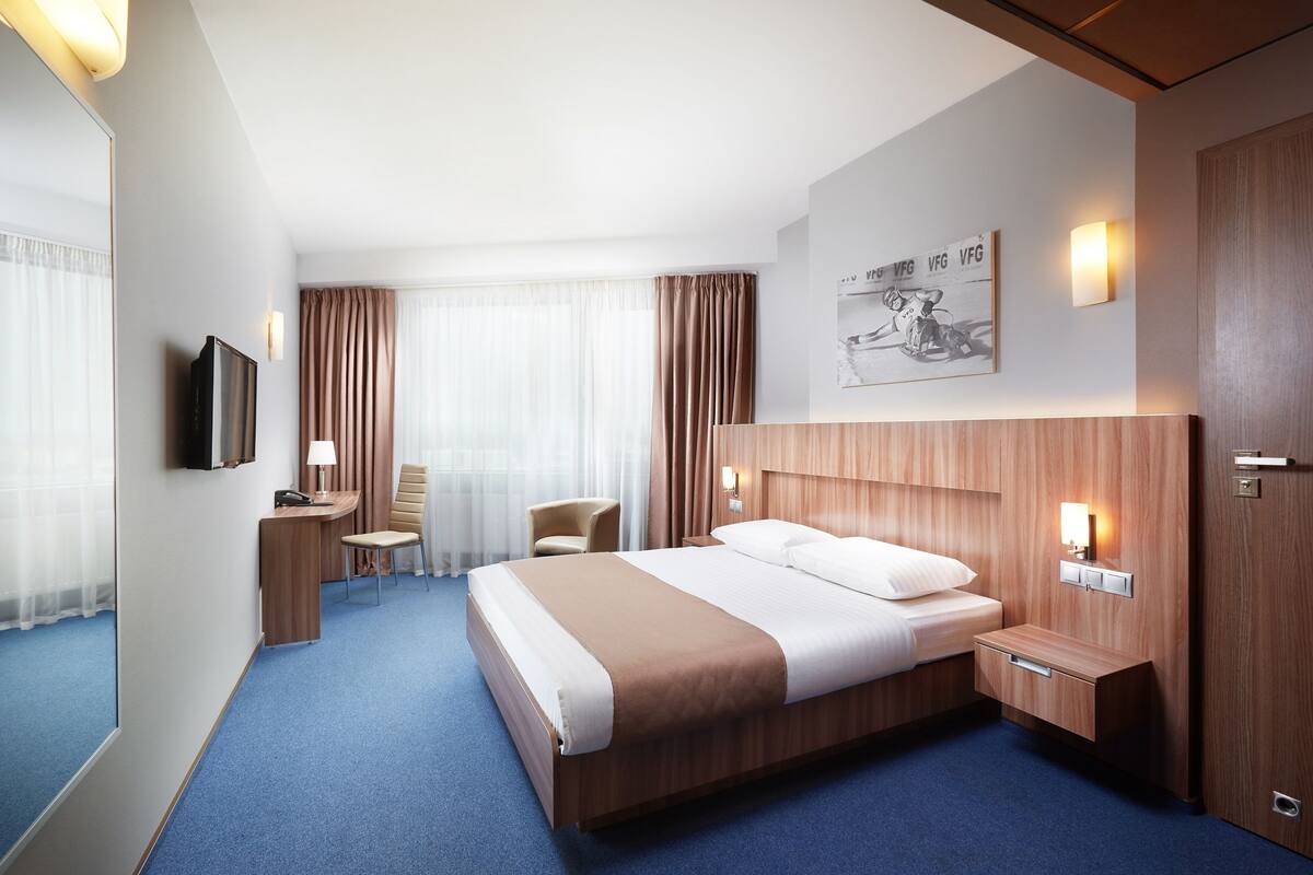 A hotel room with a large bed, a desk, a chair, a TV, and a painting on the wall.