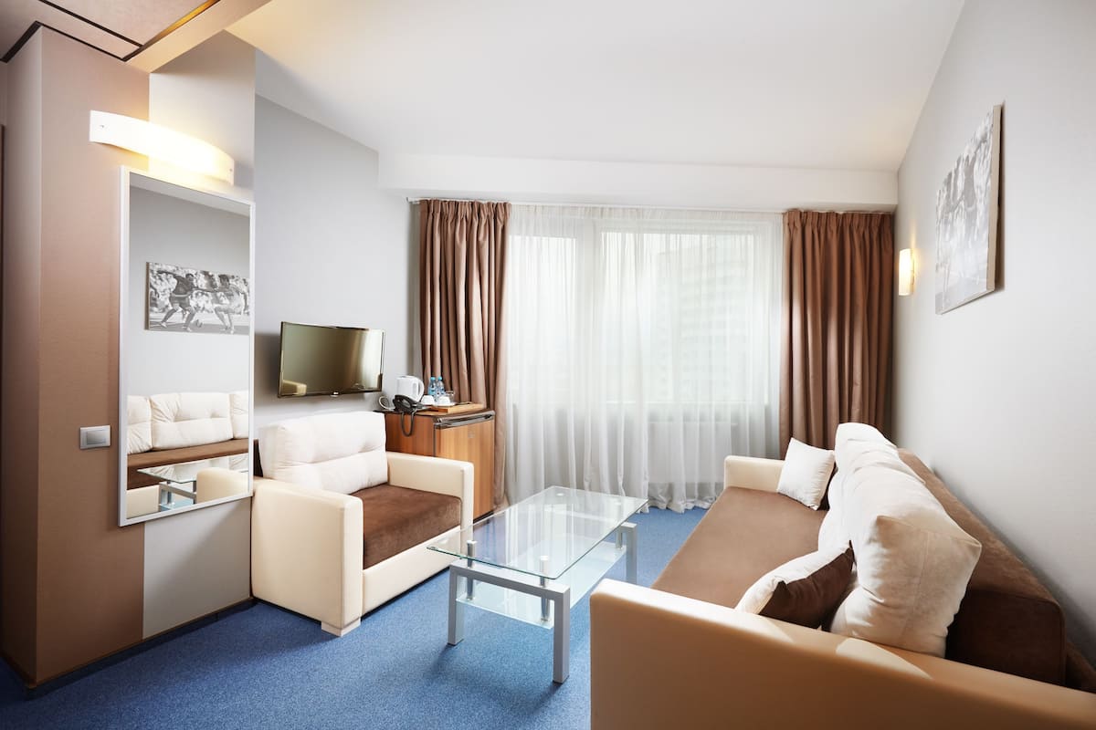 A modern hotel room with a glass coffee table, two sofas, a flat-screen TV, and a large window with curtains.