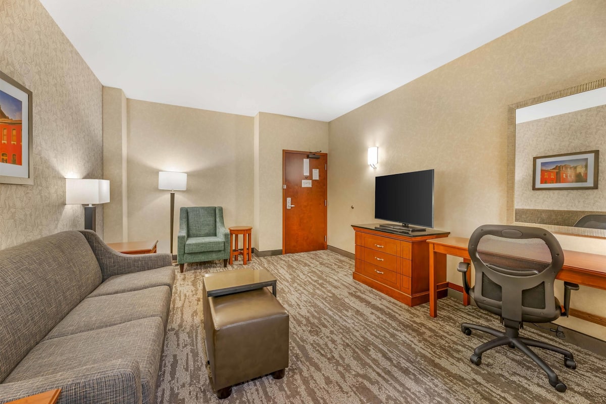 Habitación de hotel con sofá, una mesita, un escritorio con computadora, televisión y una silla.