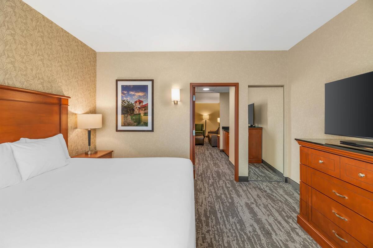 Habitación de hotel con una cama grande, un escritorio con lámpara, un televisor en un tocador y un pasillo que conduce a un baño.