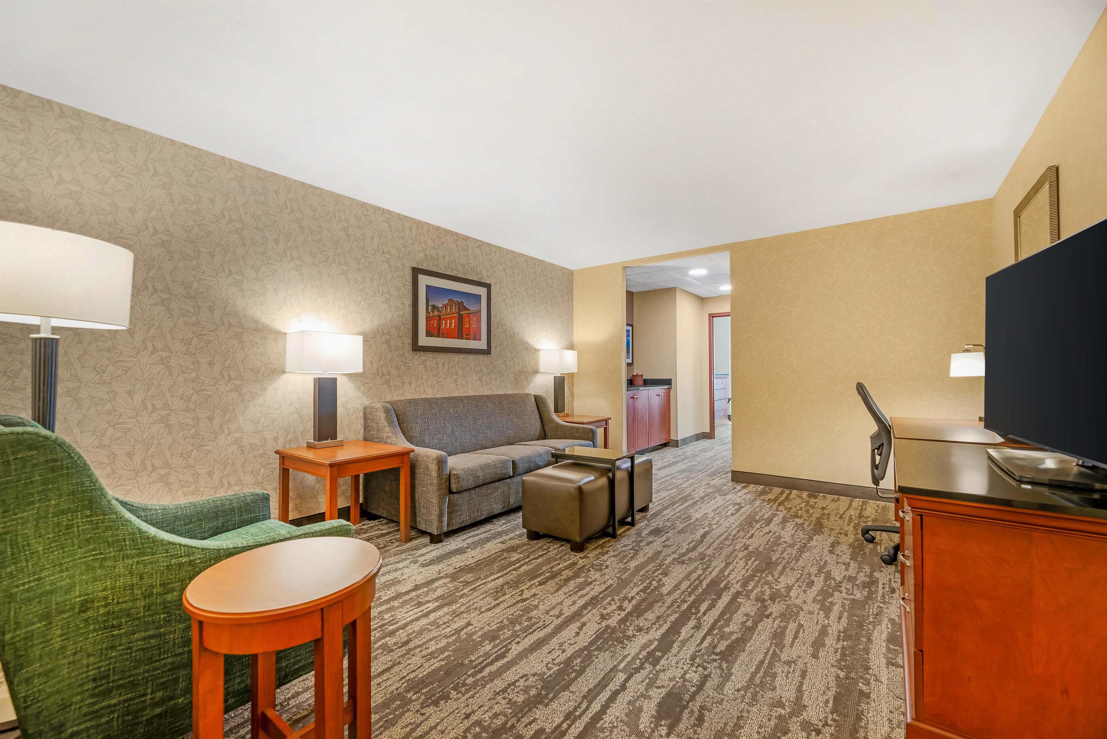 A hotel room with a green armchair, a sofa, a small table, a TV, and a desk with a chair.