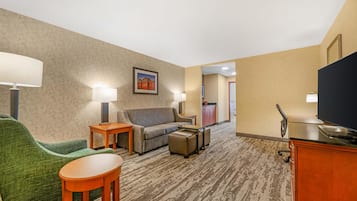 A hotel room with a green armchair, a sofa, a small table, a TV, and a desk with a chair.