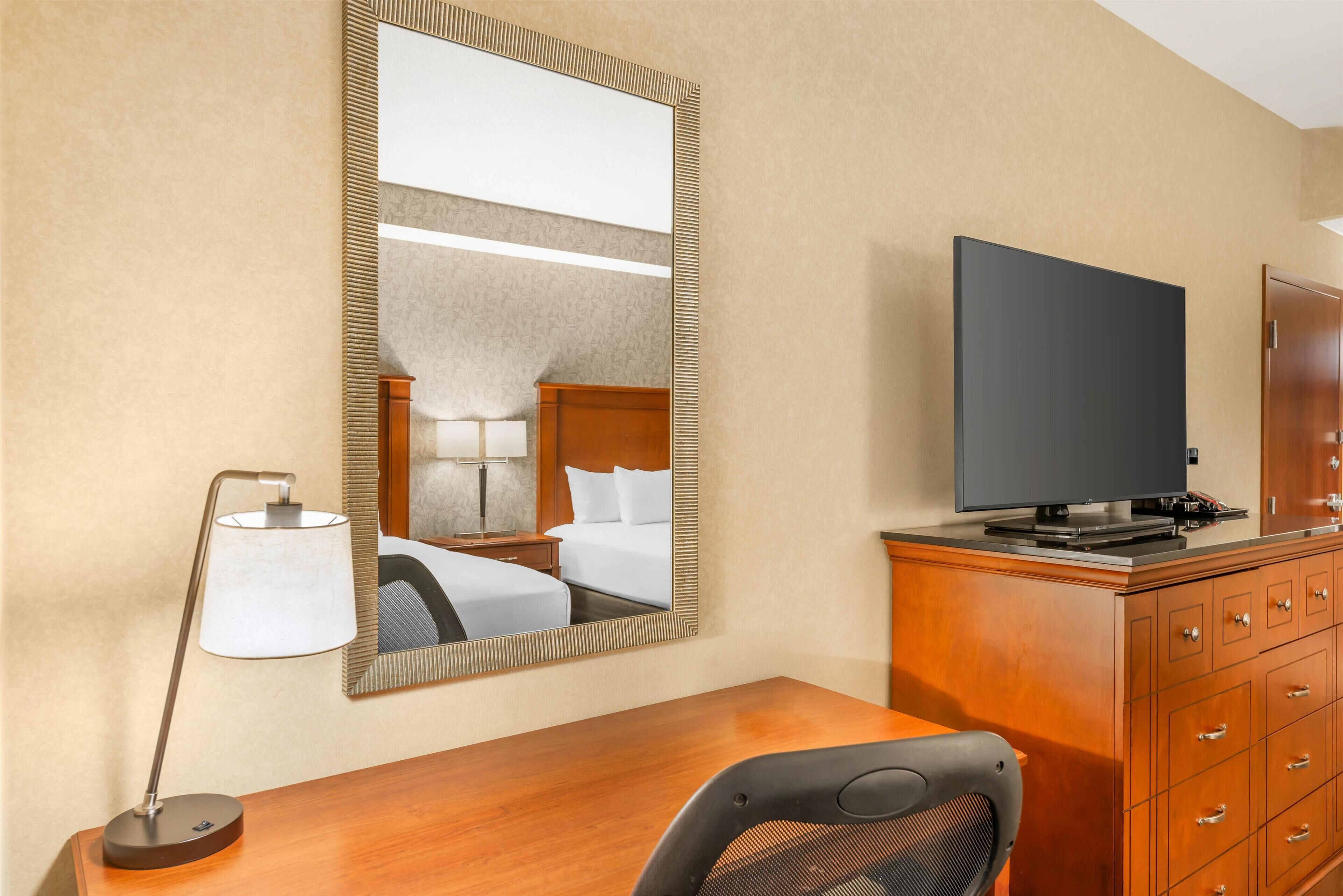 A hotel room with a wooden desk, a television on a cabinet, a large mirror, and a lamp.