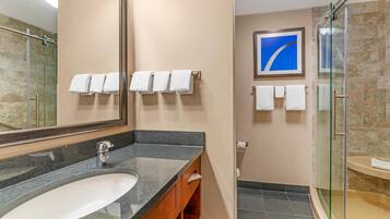 Un baño moderno con lavamanos de granito, cabina de ducha con mampara de vidrio y varios toallas blancas en toalleros.