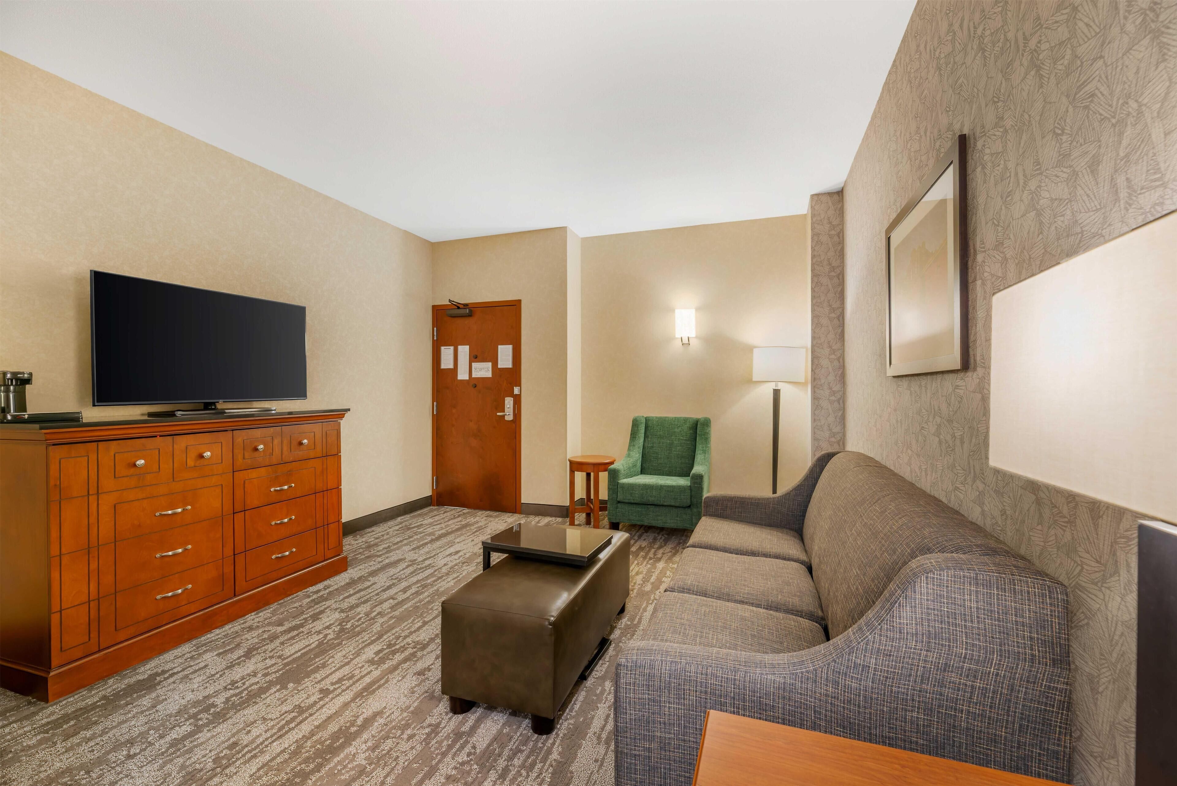 Una habitación de hotel con televisión de pantalla plana, cómoda de madera, sillón verde, sofá gris y una pequeña mesa redonda.