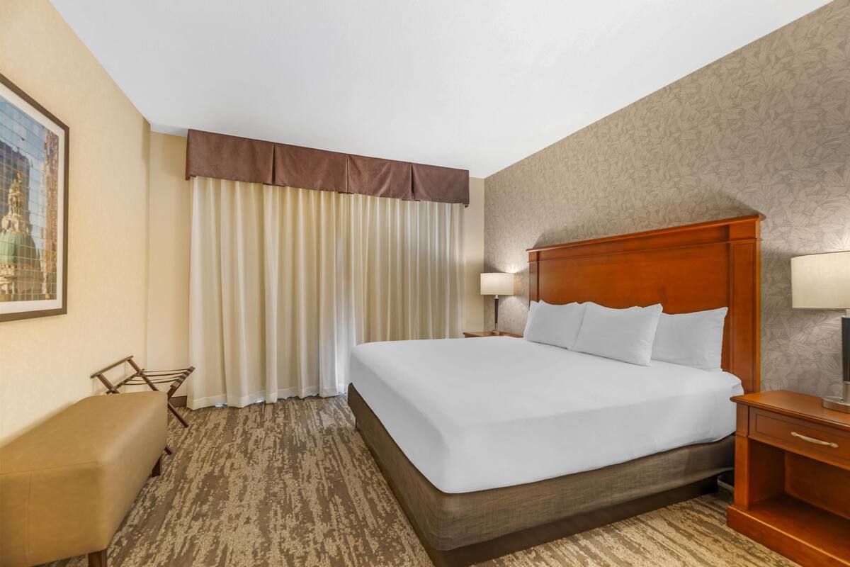 Una habitación de hotel con una cama grande, un cabecero de madera, una mesita de noche con lámpara, un banco y una ventana con cortinas.