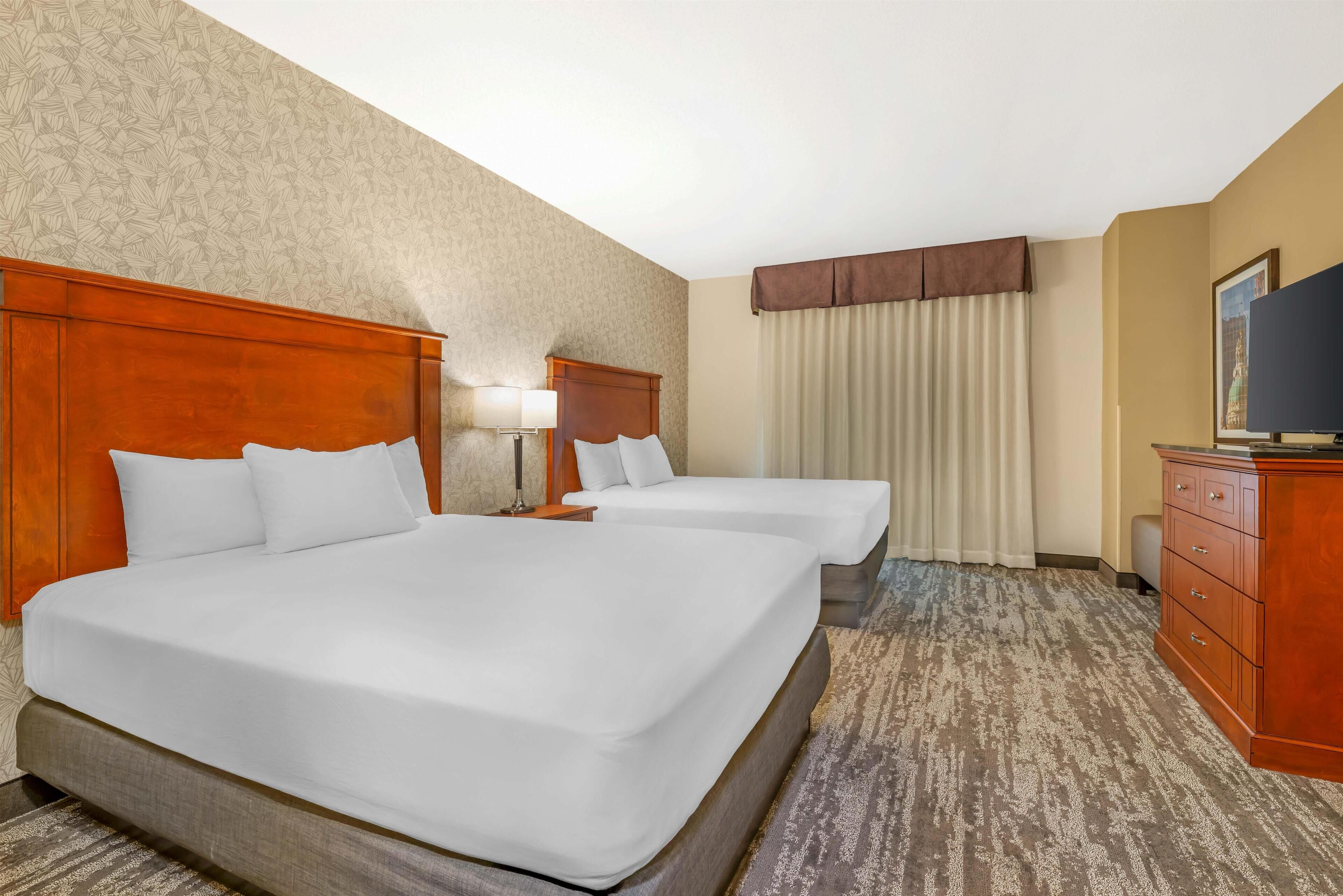 A hotel room with two beds, a wooden headboard, a flat-screen TV, a dresser, and a window with curtains.
