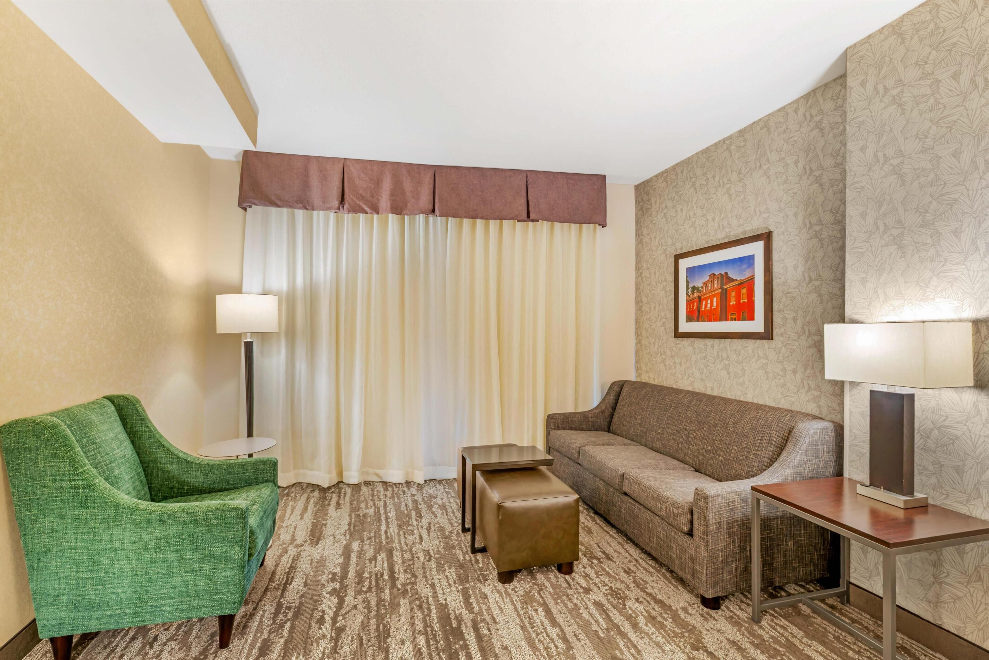A hotel room with a green armchair, a brown sofa, a small ottoman, and a side table with a lamp.
