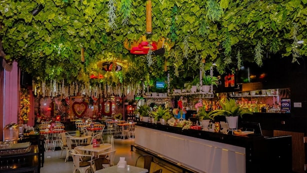 A restaurant with a green leafy ceiling, white tables, and a bar area with a variety of bottles and decorations. - G Suites Hotel by AMITHYA (Surabaya)