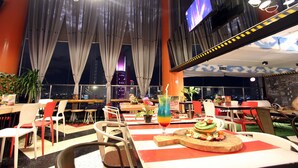 A restaurant with tables, chairs, and a large TV screen. - G Suites Hotel by AMITHYA (Surabaya)