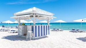 Eine Strandbar mit weiß-blauen Streifen, weißen Sonnenschirmen und Liegestühlen direkt am Sandstrand, mit dem Meer im Hintergrund.