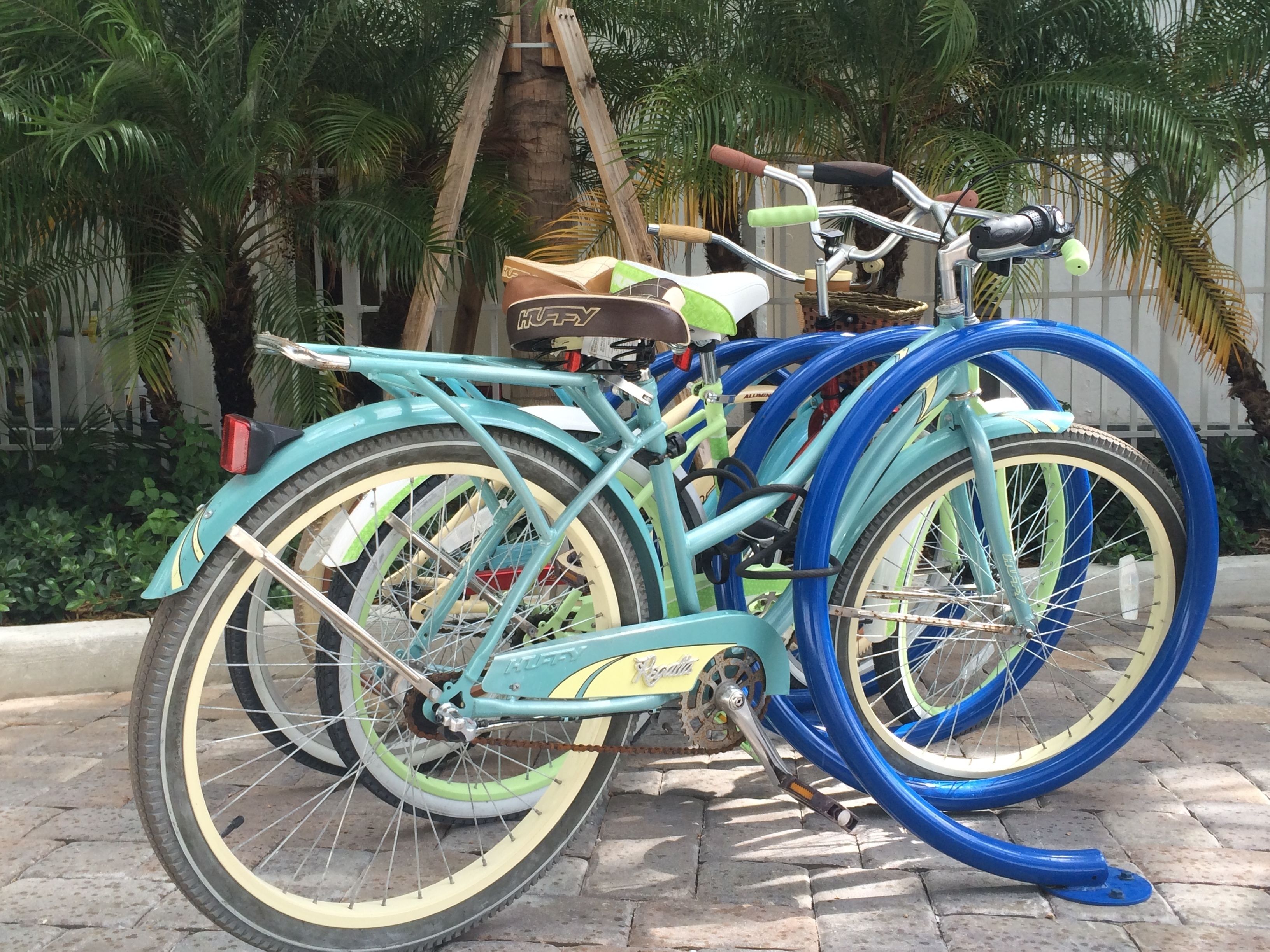 Una bicicleta turquesa con asiento verde y detalles amarillos, estacionada sobre una superficie pavimentada con palmeras al fondo.