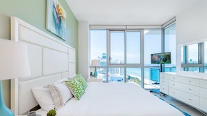 Ein modernes Hotelzimmer mit einem großen Bett, einem Fernseher, einem Schminktisch und Blick auf die Stadt durch große Fenster.