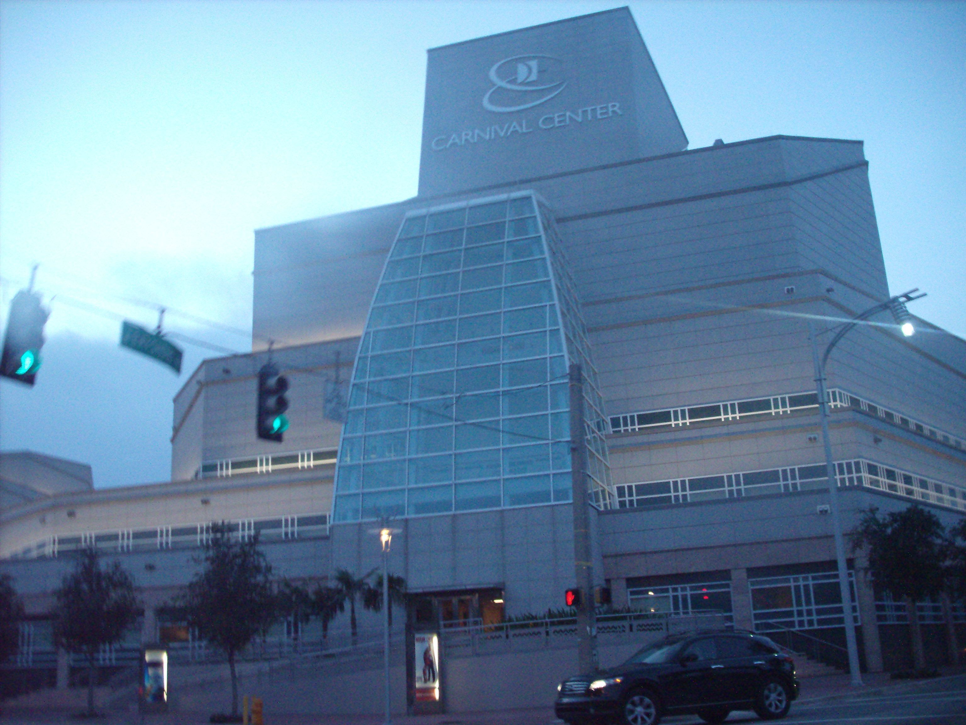 Un bâtiment moderne doté d’une façade distinctive en verre et d’une grande structure angulaire sur le toit, portant l’inscription « Carnival Center ».