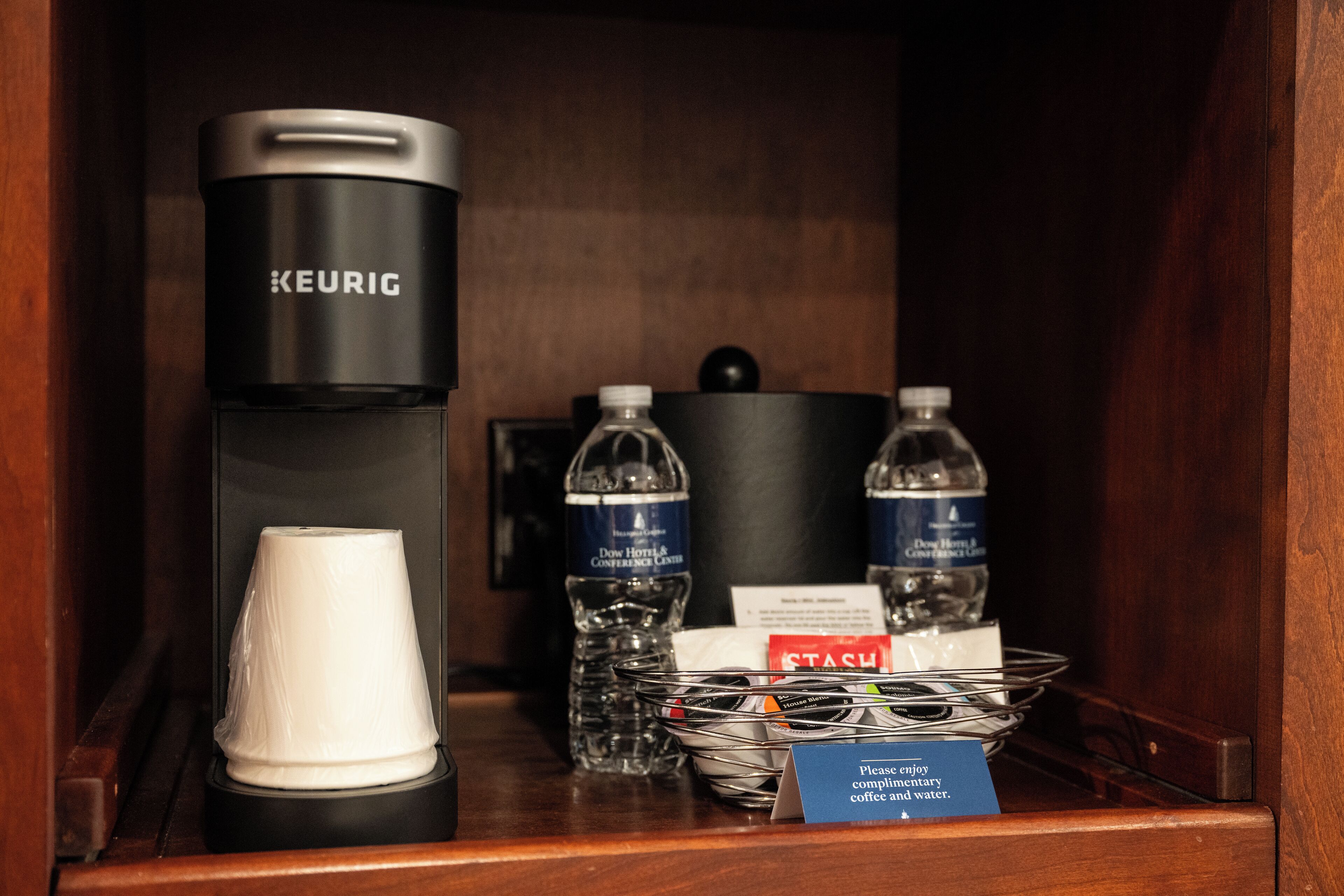 A coffee station with a Keurig coffee maker, disposable cups, water bottles, and a "Please enjoy complimentary coffee and water" sign.