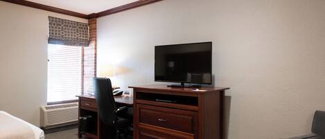 A hotel room with a bed, a desk with a chair, a television, and a window with blinds.