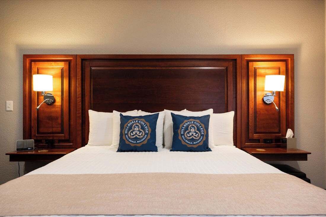 Habitación de hotel con una cama grande, dos almohadas con un logotipo, mesitas de noche con lámparas y una pequeña mesa con lámpara.