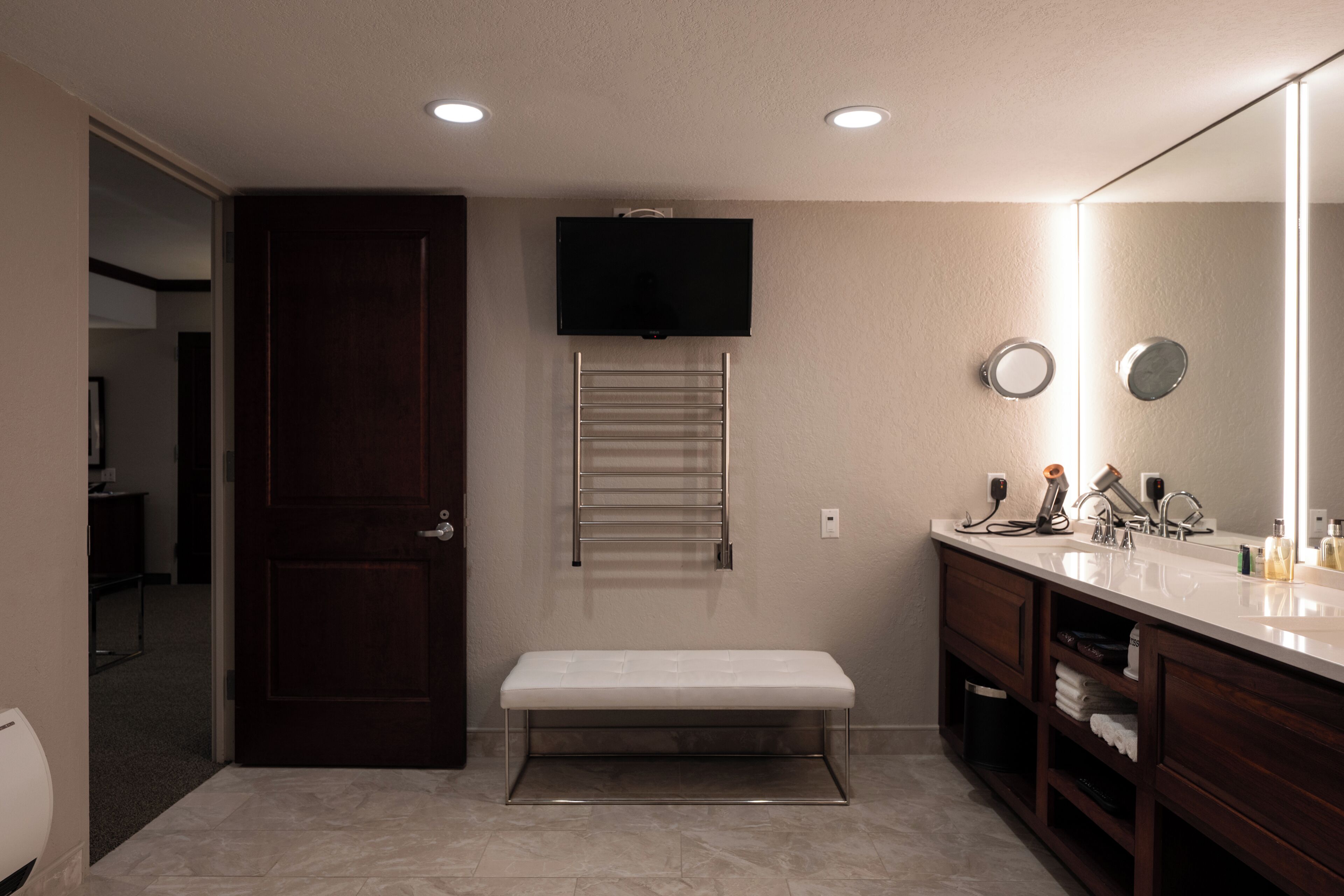 A bathroom with a bench, a towel rack, a mirror, and a television mounted on the wall.
