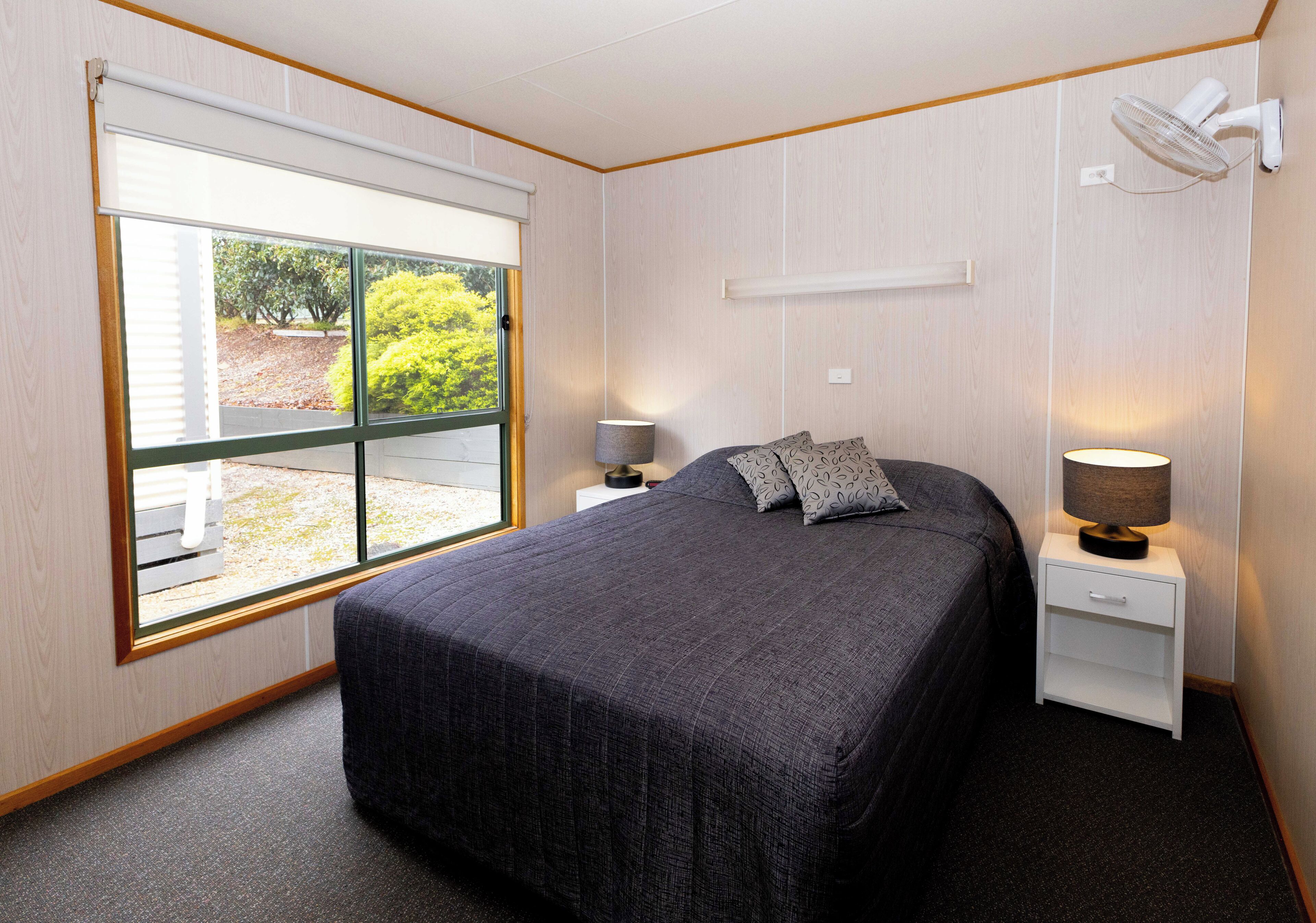 A bedroom with a large bed, two bedside tables with lamps, a window with a view of greenery, and a ceiling fan.