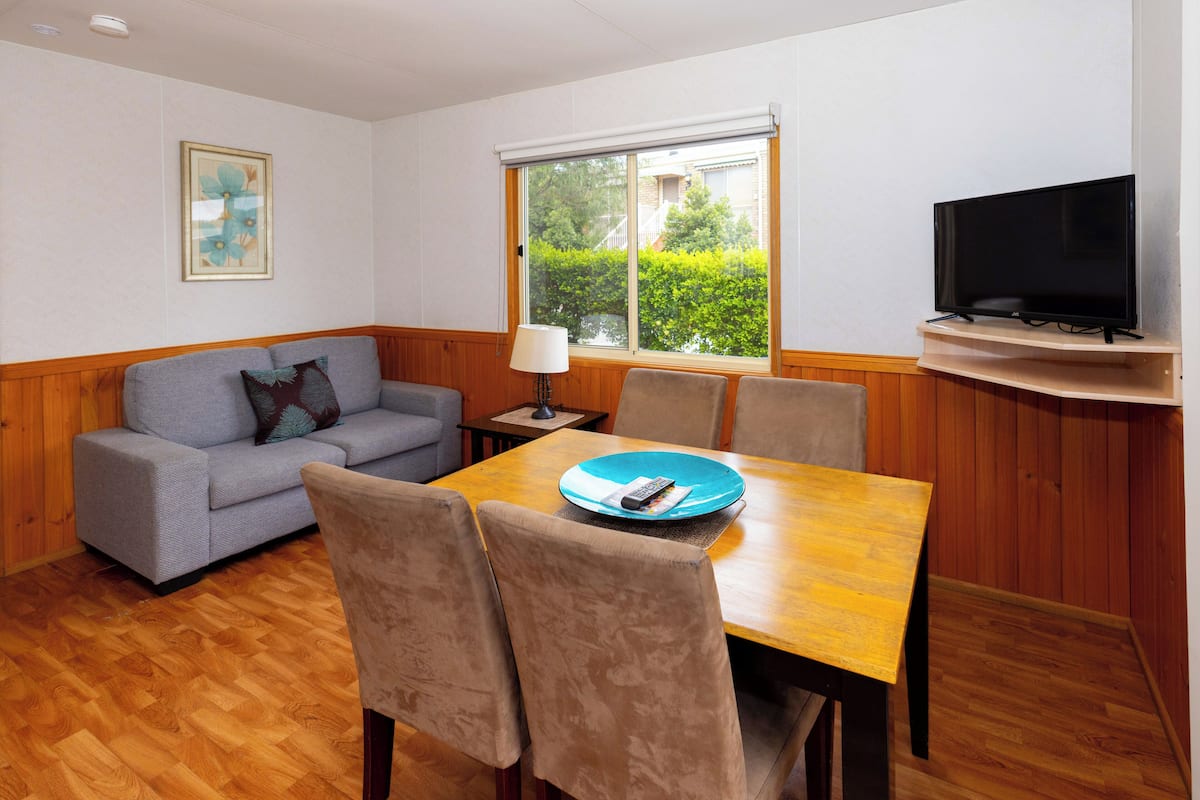 Un salón con sofá, sillas, mesa, televisor y ventana con vistas a follaje.