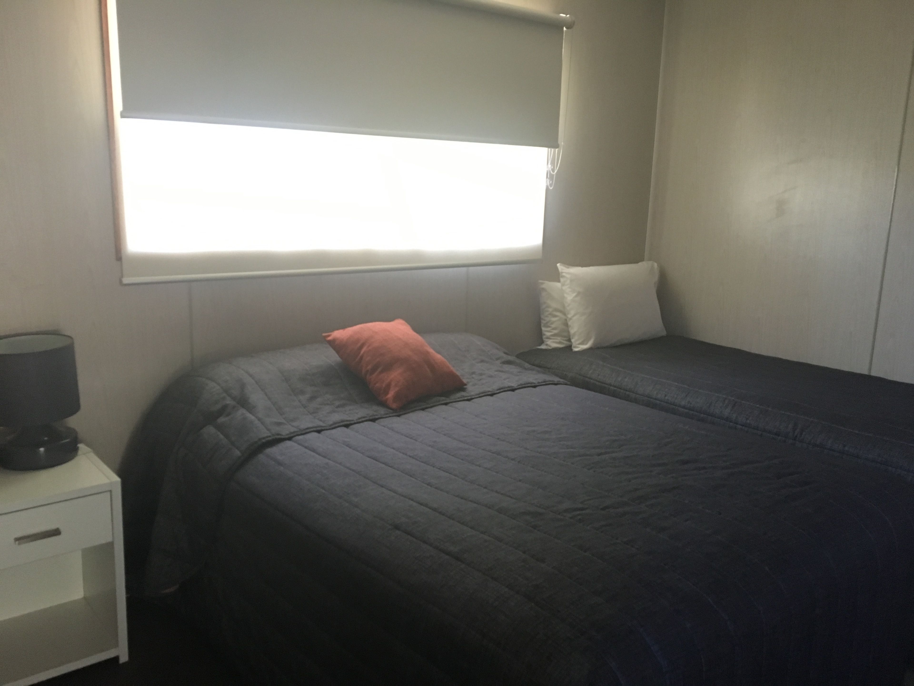 Una cama individual con una manta oscura y un cojín rojo, una mesita de noche con una lámpara y una ventana con una persiana blanca.