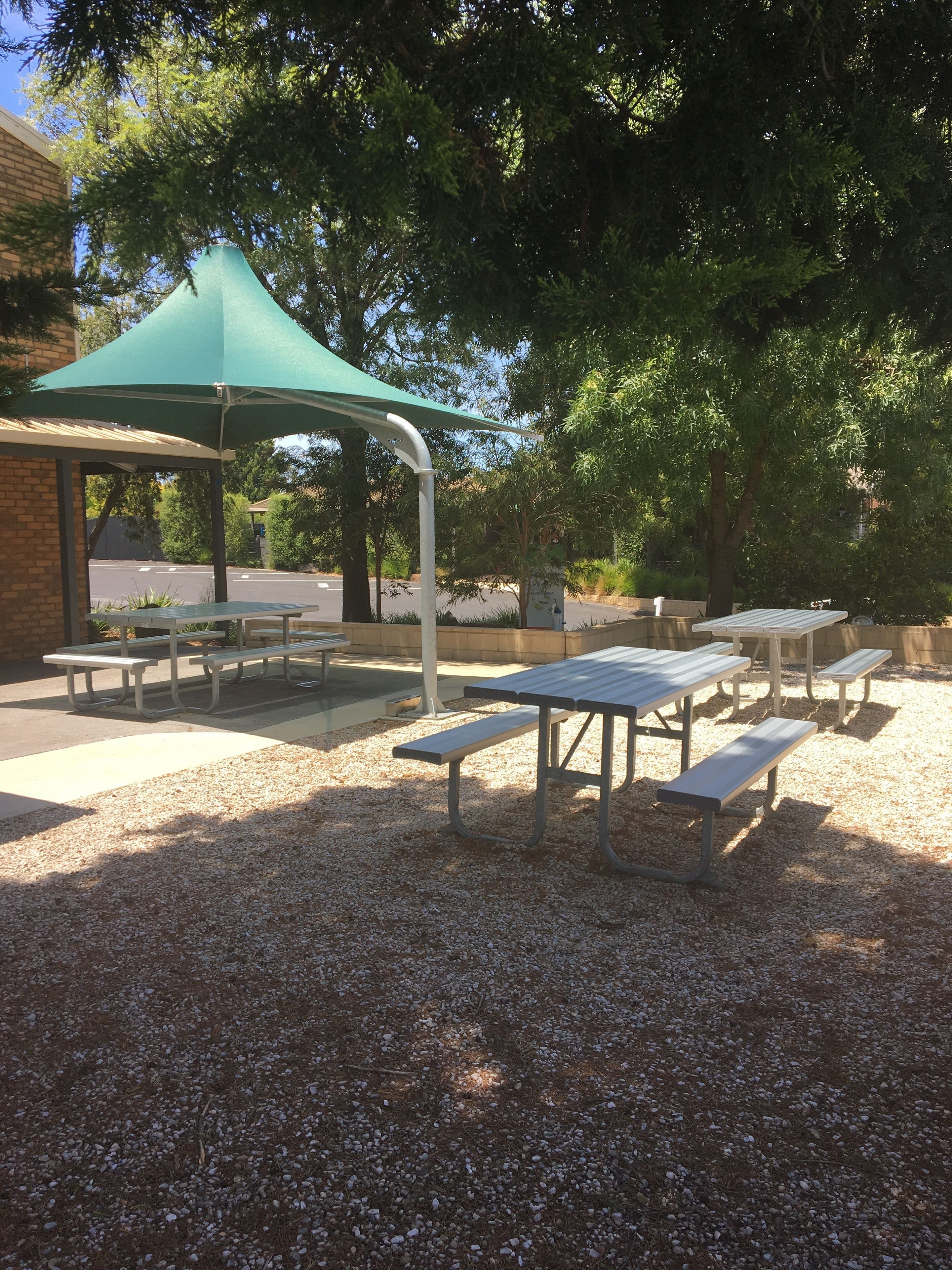 Un espace pique-nique abrité, équipé de tables et de bancs, entouré de gravier et d’arbres.