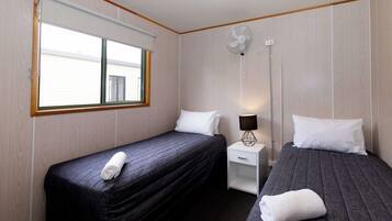 A hotel room with two single beds, a window with blinds, a bedside table with a lamp, and a wall-mounted fan.