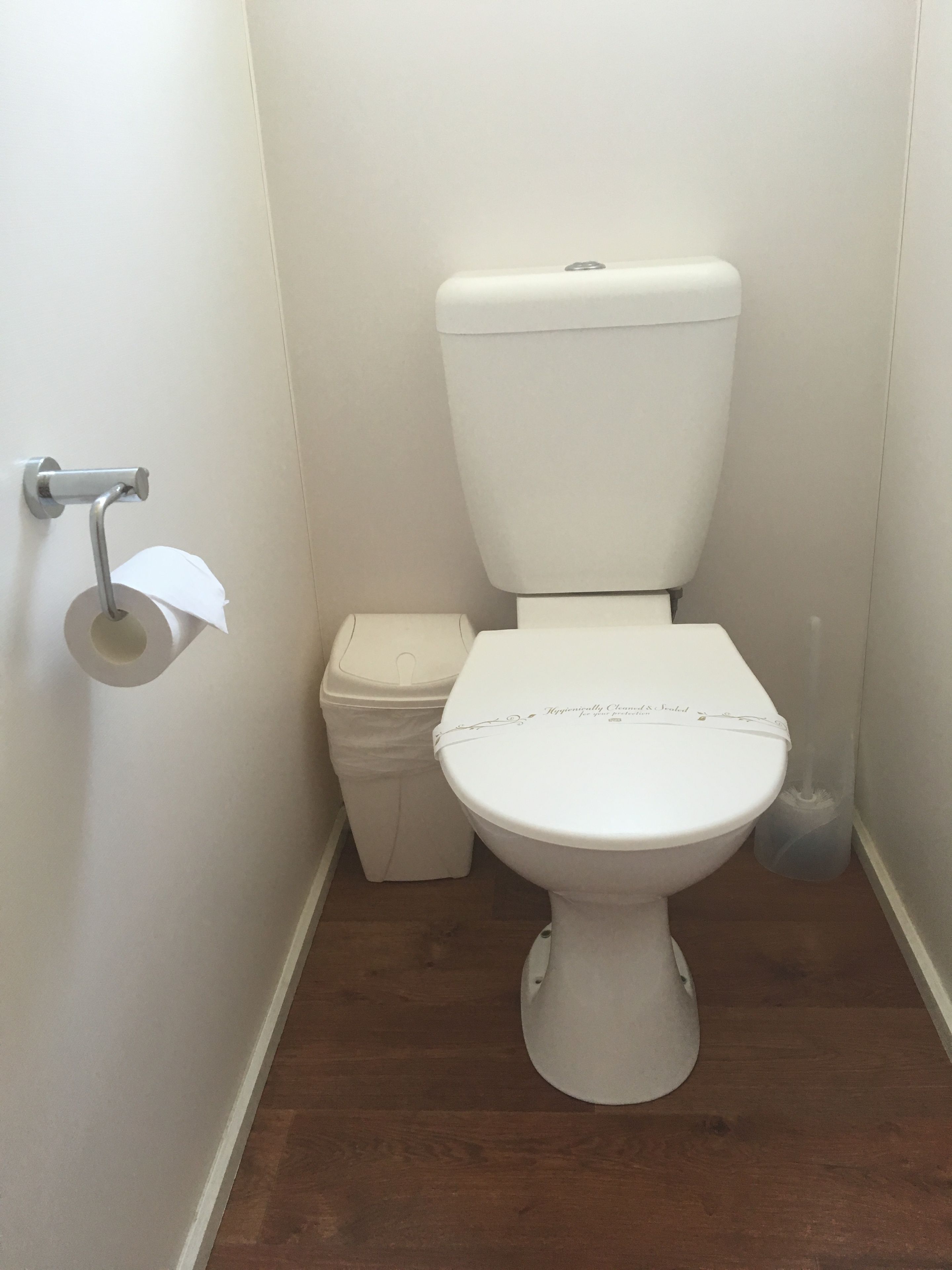 Des toilettes blanches avec un abattant, un porte-rouleau de papier toilette et une poubelle, dans une salle de bain dont le sol est recouvert de parquet.