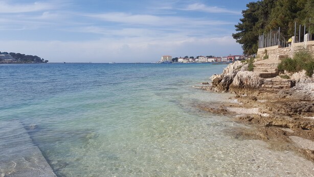 A coastal scene with clear shallow waters, rocky shoreline, and buildings in the background. - Hotel Villa Karda (Porec)