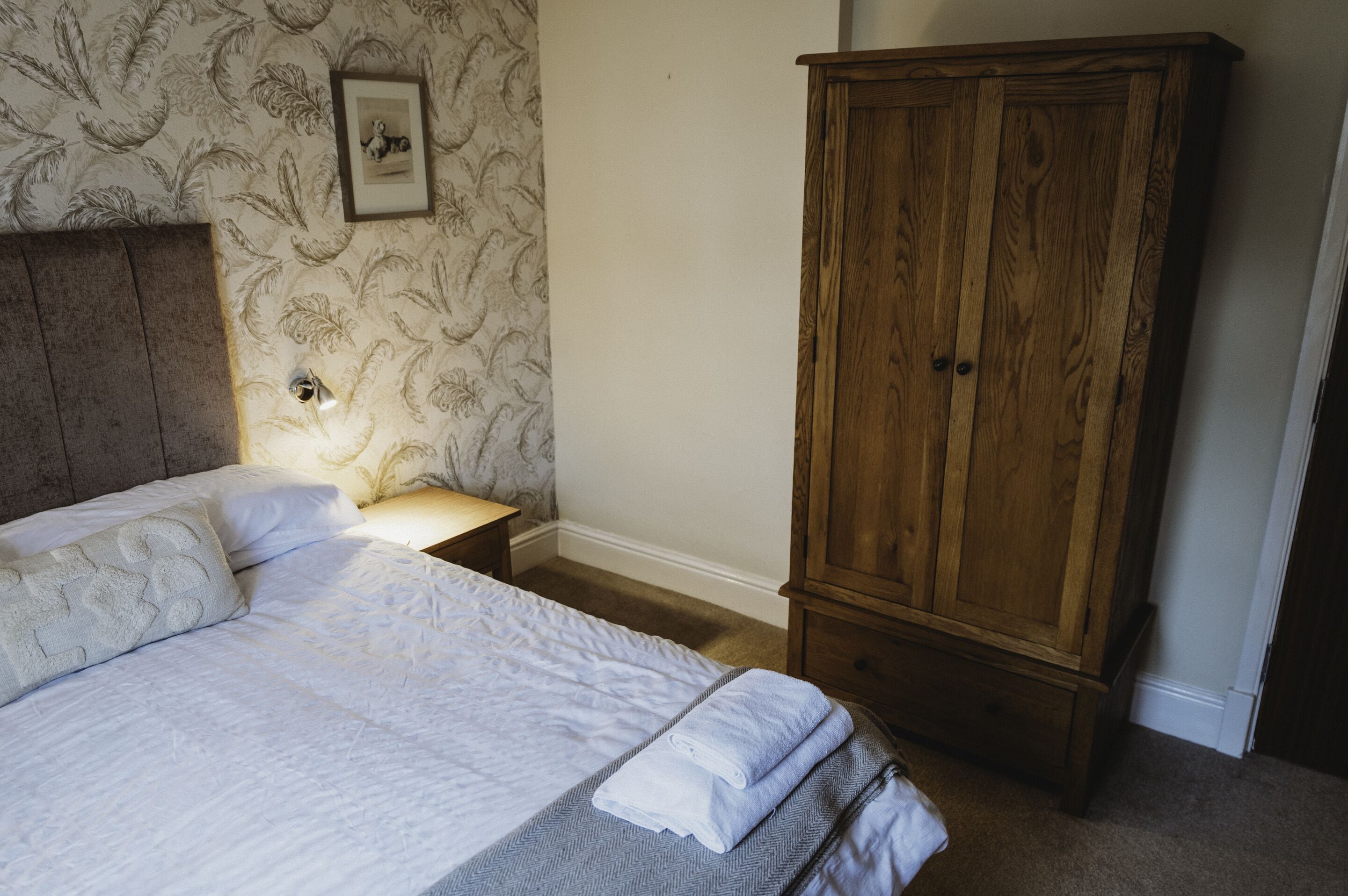 Un dormitorio con una cama, una mesita de noche, un armario de madera y un papel tapiz con motivos florales.