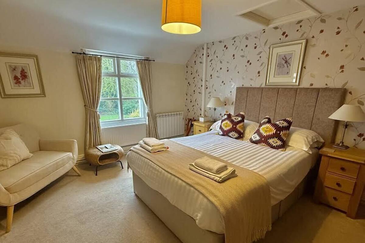 A bedroom with a large bed, a bedside table, a nightstand, a chair, a window with curtains, and a wall with floral wallpaper.