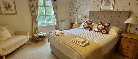 Une chambre à coucher avec un grand lit, une table de chevet, une commode, une chaise, une fenêtre avec des rideaux et un mur recouvert d’un papier peint à motifs floraux.