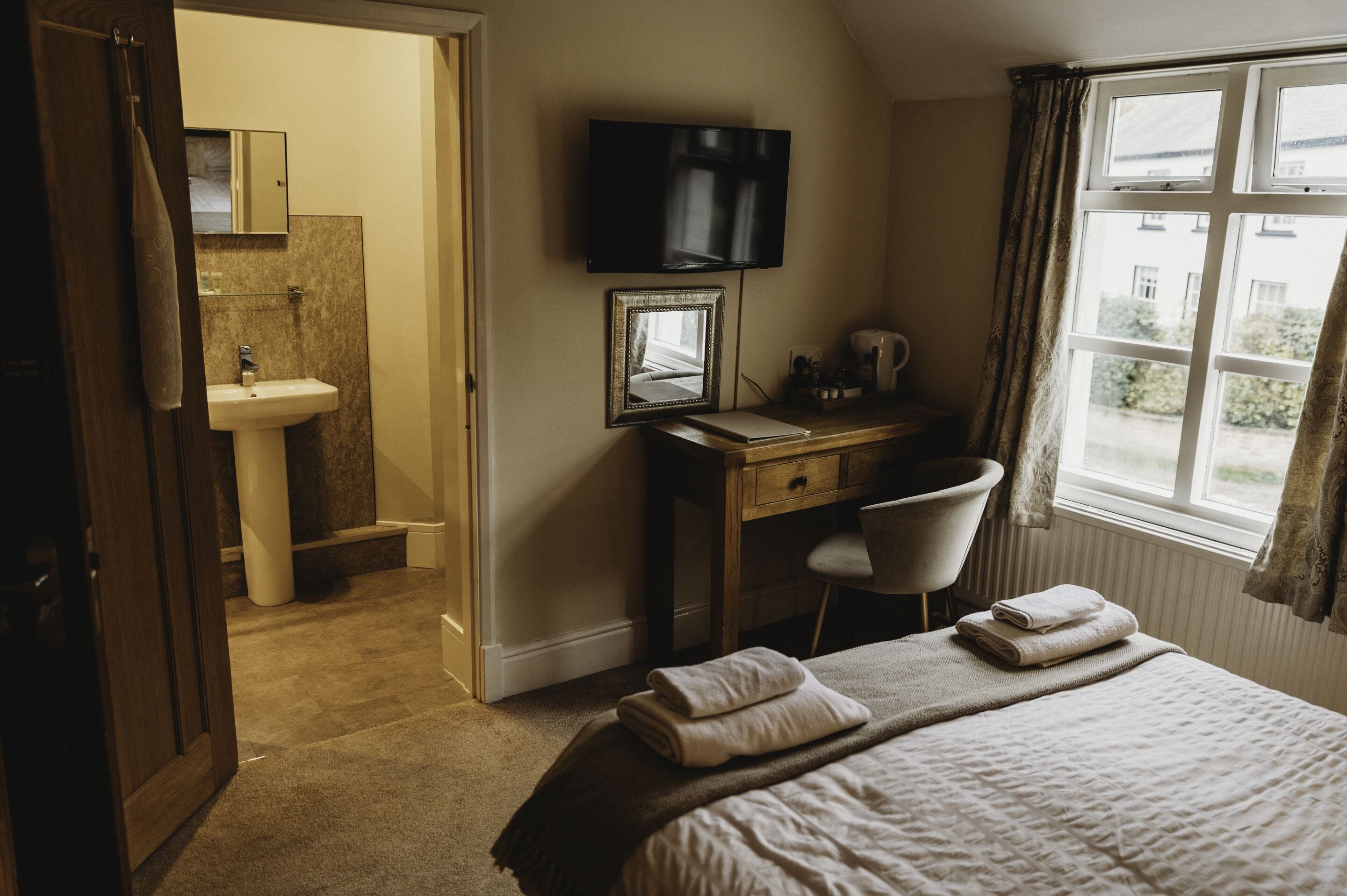 A hotel room with a bed, a desk with a chair, a television, a bathroom with a sink, and a window with curtains.