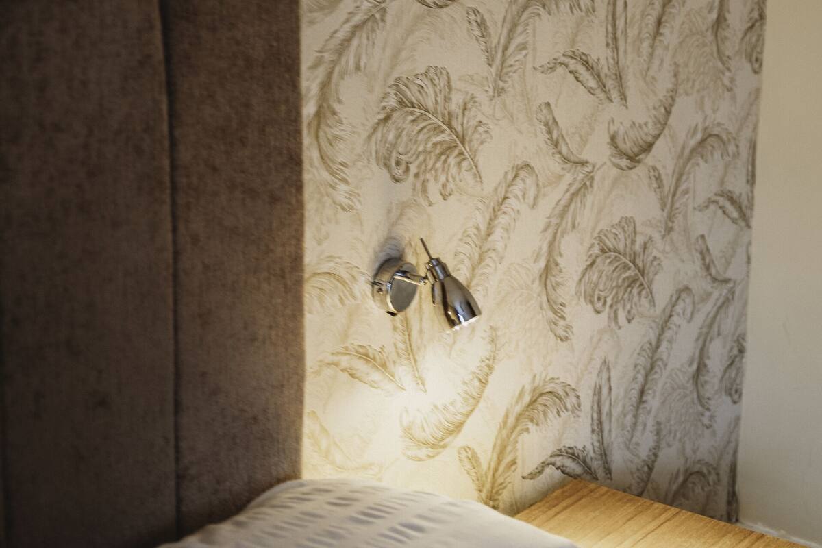 A bedroom with a headboard, a bed with white bedding, a nightstand, a framed picture on the wall, and a wall-mounted lamp.