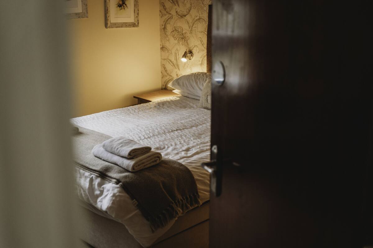 A neatly made bed with a folded towel and a blanket, a bedside lamp, and a wall with a floral pattern.