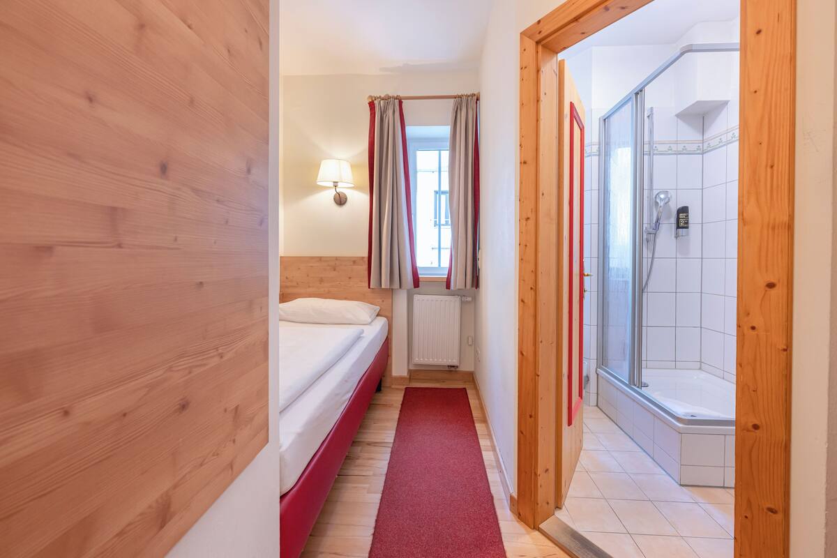 Camera d'albergo stretta con pavimento in legno, un letto, un tappeto rosso, un angolo doccia e una finestra con tende.