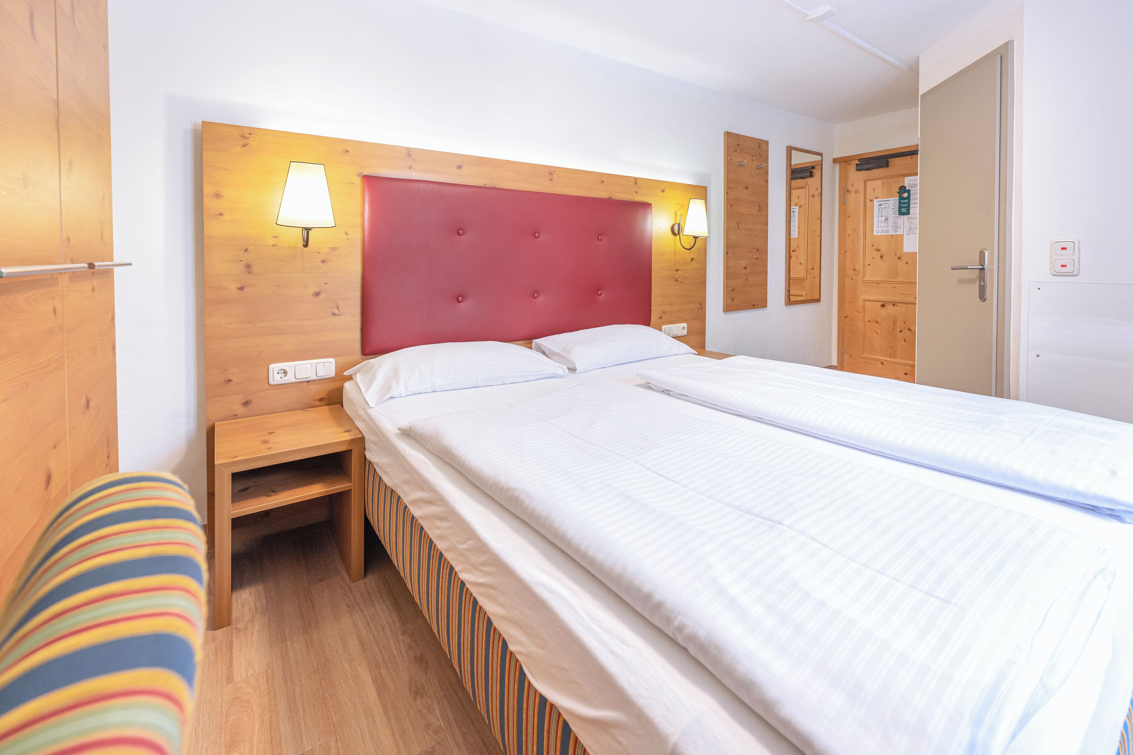 Una cama doble con cabecera roja, mesitas de noche de madera y un banco a rayas.