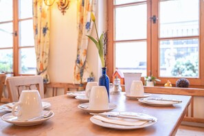 Ein Esstisch mit weißen Tellern, Tassen und Besteck, eine blaue Vase mit Pflanze und im Hintergrund ein Fenster mit Vorhängen.