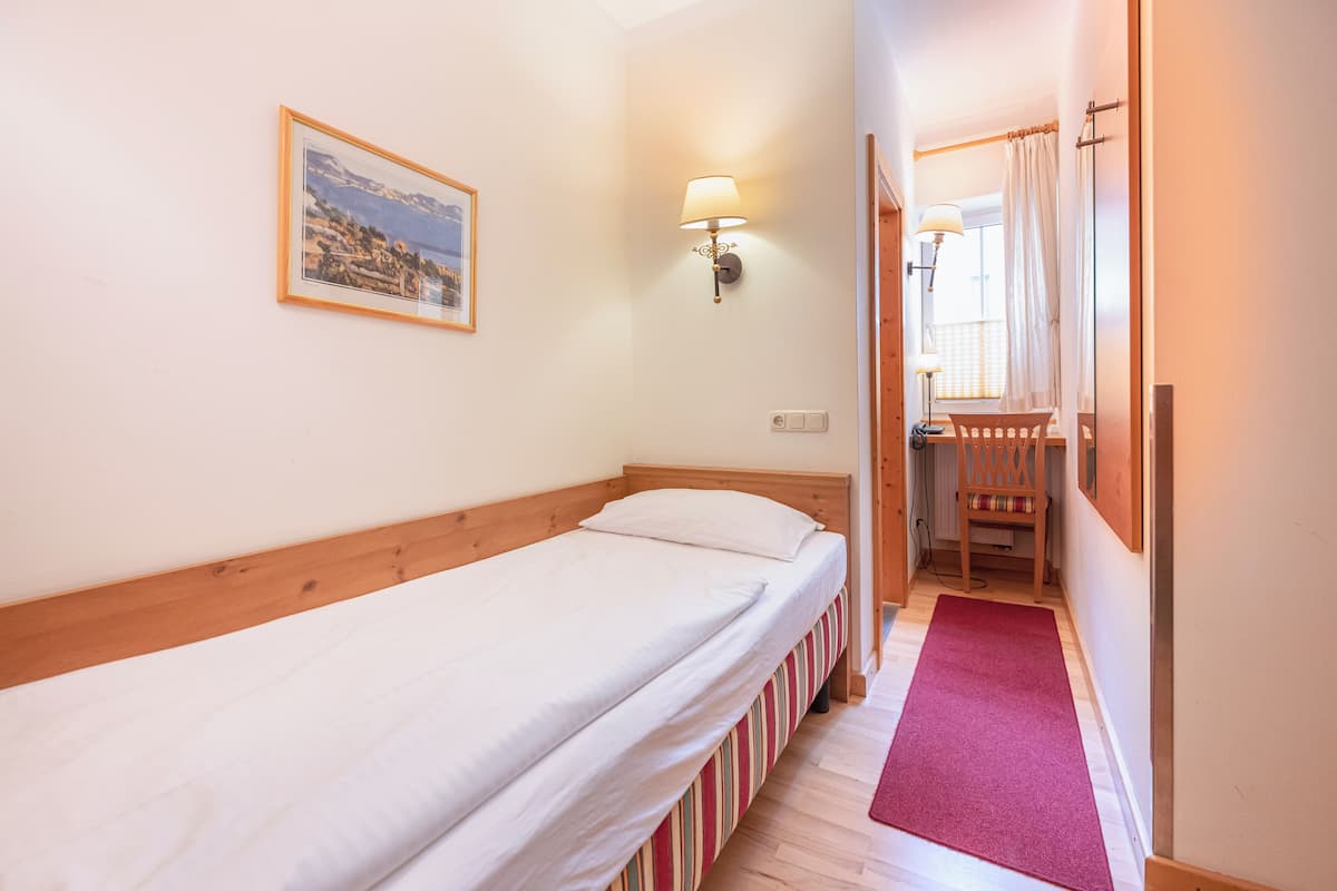 Un letto singolo con testiera in legno, un comodino, una lampada fissata al muro, un quadro incorniciato e un tappeto rosso che conduce a un balcone.
