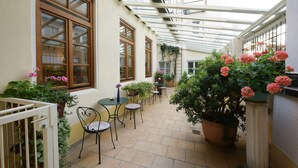 A covered patio with tables and chairs, potted plants, and a view of the street through large windows. - Eder  (Munich)
