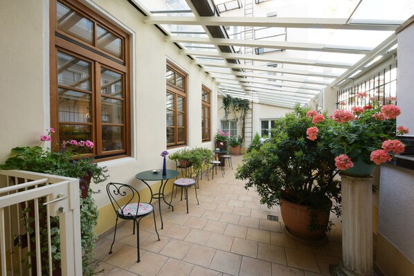 Eine überdachte Terrasse mit Tischen und Stühlen, Blumentöpfen und Blick auf die Straße durch große Fenster.