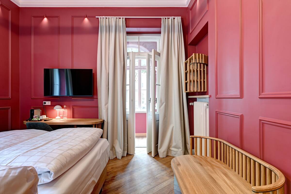 A hotel room with two beds, a TV, a desk with a lamp, and a wooden bench.