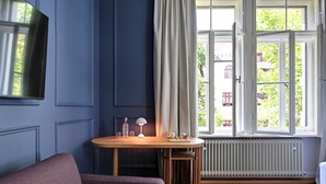 A room with a blue wall, a wooden table with a lamp, a small stool, and a radiator. - Hotel Uhland (Munich)