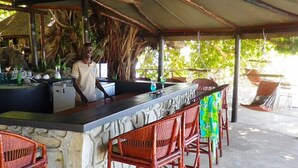 Zona de bar al aire libre con techo, barra de piedra, sillas de madera y una persona de pie detrás de la barra.