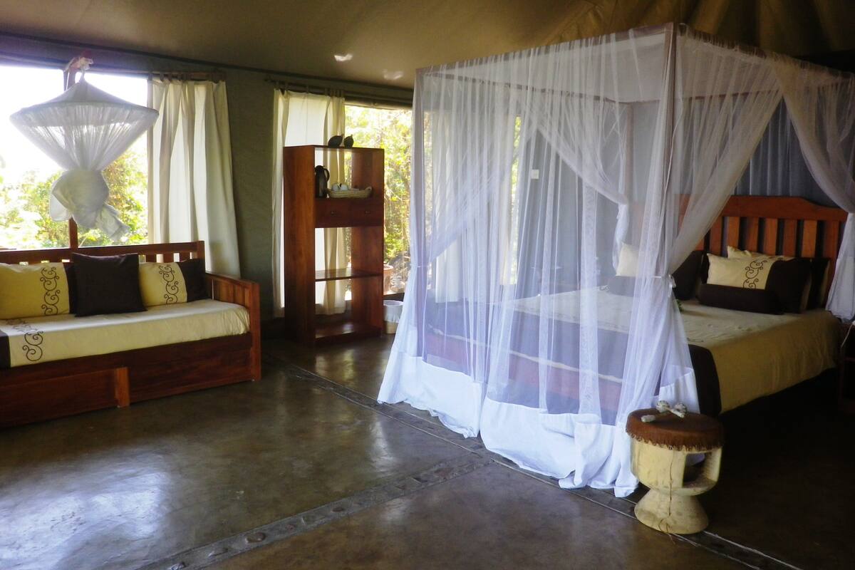 A room with a bed under a canopy, a sofa with cushions, a wooden cabinet, and a small stool.