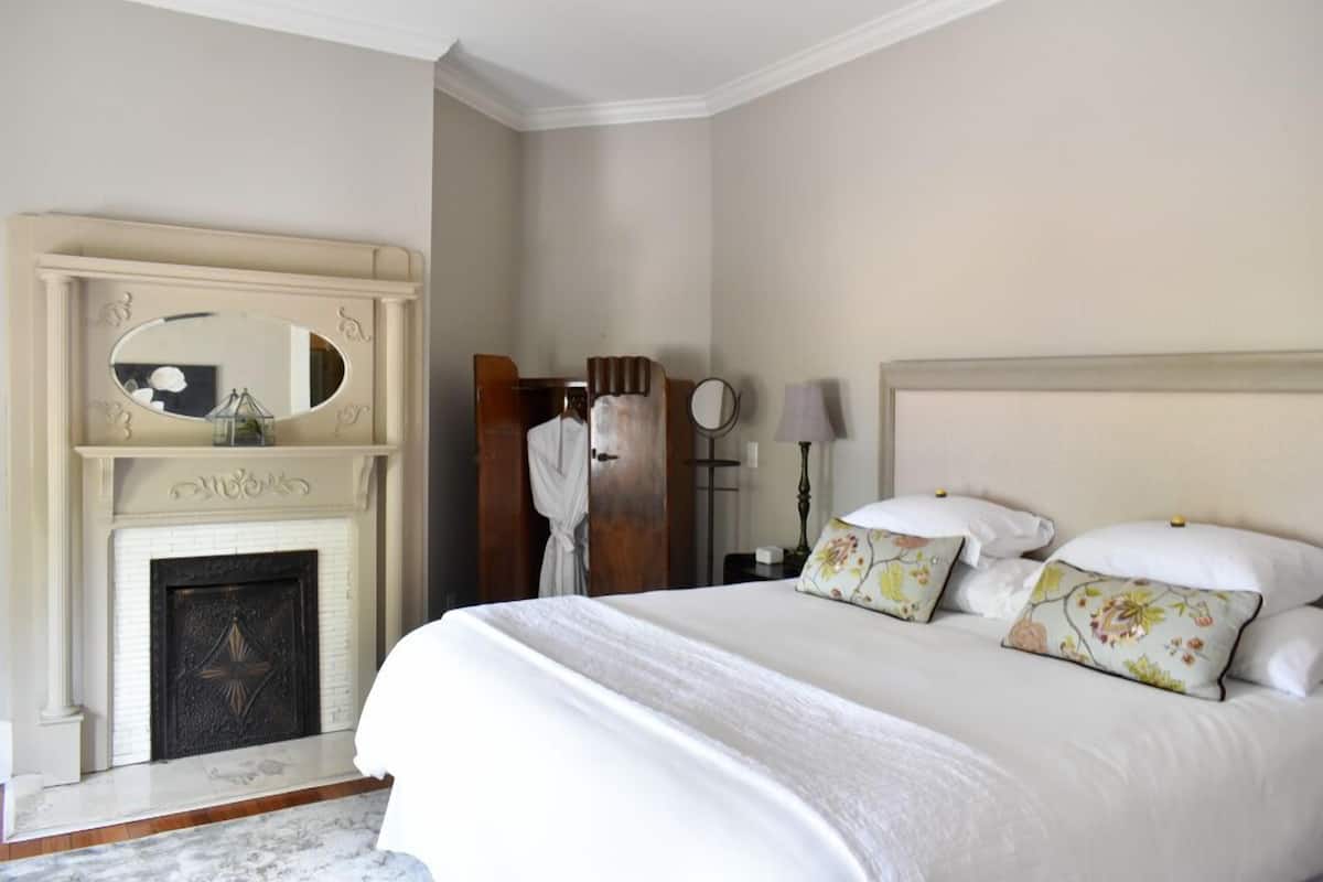 A bedroom with a large bed, a fireplace, a mirror, and a nightstand with a lamp.